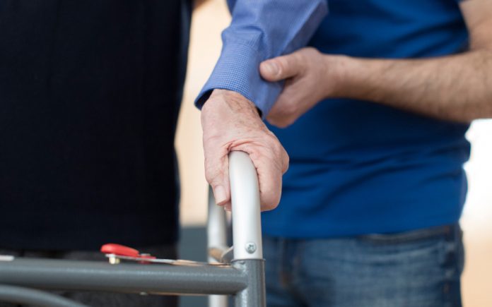 Senior Man's Hands On Walking Frame With Care Worker In Background