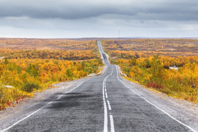 Northern country road among hills with colorful autumn tundra trees and bushes on a cloudy day. Kola Peninsula, Murmansk region, Russia