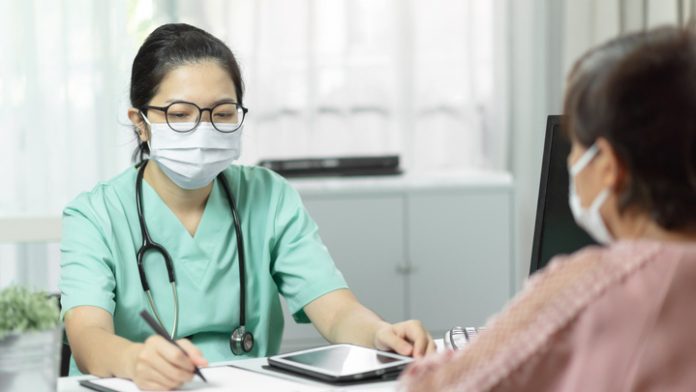 Asian female Doctor in green uniform wear eyeglasses and surgical mask writing something in checklist document while talking with Elderly woman patient in medical room at the hospital. Symptoms, Disease, Flu. Covid-19, Coronavirus