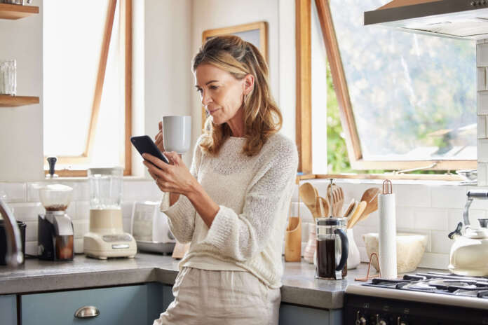 Coffee, phone and senior woman in kitchen browsing social media, text message or online app at home. Relax, tea and reading news or mobile online web surfing with female from Canada on 5g smartphone