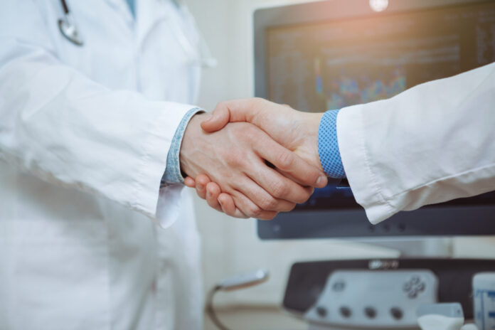 Cropped close up of two male doctors in labcoats shaking hands at the hospital, celebrating success. Male practitiner shaking hands with colleague at work. Medical team, achievement, science concept